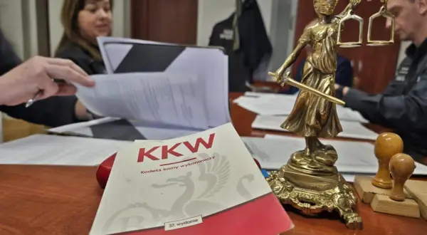 Na biurku leży Kodeks karny wykonawczy, obok Statua Temidy. W tle widać osoby pracujące nad dokumentami, co może sugerować sprawy związane z dozorem elektronicznym.