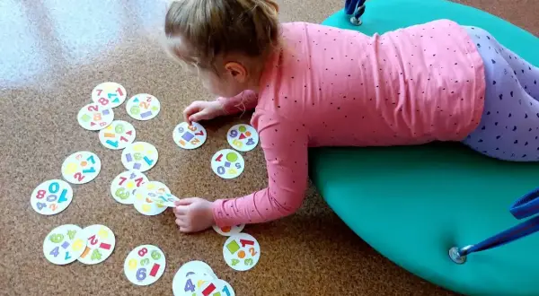 Dziewczynka na huśtawce bawi się kartami z cyframi i kształtami. To element jej rewalidacji, wspierający rozwój.