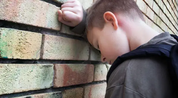 Chłopiec z plecakiem opiera głowę o ceglaną ścianę, wyrażając smutek i poczucie osamotnienia, które mogą być objawami fobii szkolnej.