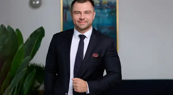 Mężczyzna w garniturze, stojący obok rośliny, z obrazem portowym w tle.