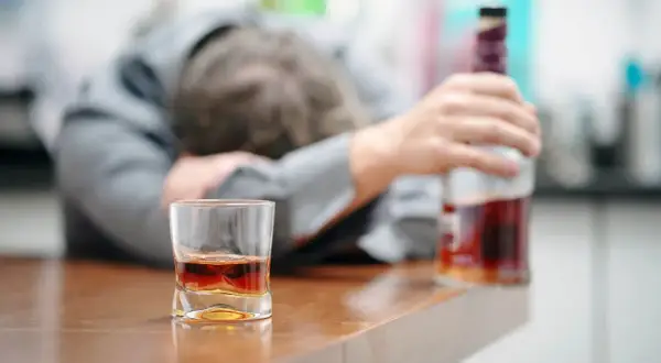 Ubezwłasnowolnienie alkoholika. Mężczyzna w garniturze leży na blacie, obok szklanka z whisky i butelka.