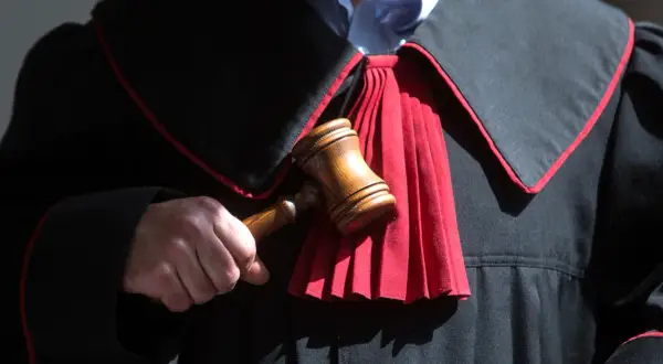 Sędzia trzyma młotek, gotowy do ogłoszenia wyroku. W tle widać togę z czerwonym żabotem, symbolizującą sprawiedliwość i powagę, która towarzyszy wydaniu subsydiarnego aktu oskarżenia.