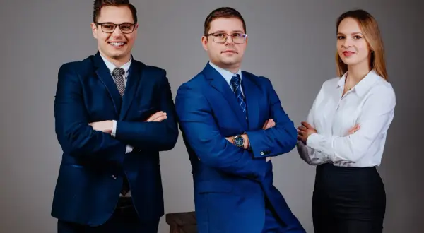 Trzy osoby w garniturach i białej koszuli, stojące z rękami skrzyżowanymi na piersiach.