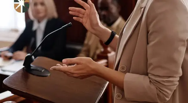Oskarżyciel posiłkowy przemawia przy mównicy, gestykulując. W tle siedzą sędziowie.