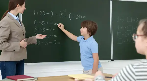 Nauczycielka pomaga uczniowi rozwiązać zadania matematyczne przy tablicy. Dzieci uczą się, wypełniając swój obowiązek szkolny.