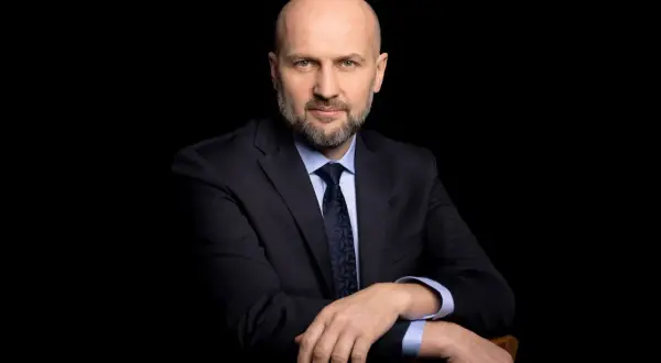 Łysy mężczyzna w garniturze i krawacie, oparty o krzesło, na czarnym tle.