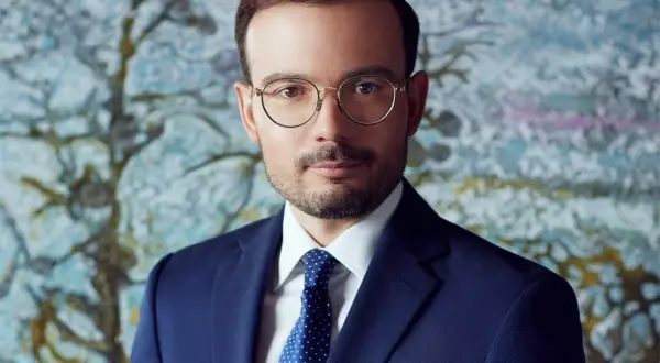 Mężczyzna w okularach i granatowym garniturze na tle abstrakcyjnego obrazu.