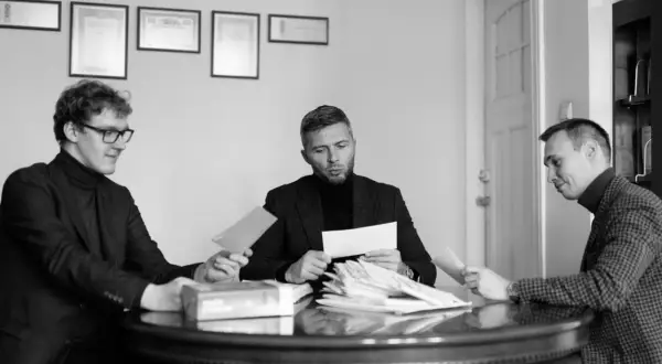 Trzech mężczyzn przy okrągłym stole analizuje dokumenty. Jeden z nich trzyma kopertę, jakby właśnie otrzymał wyrok w zawieszeniu.