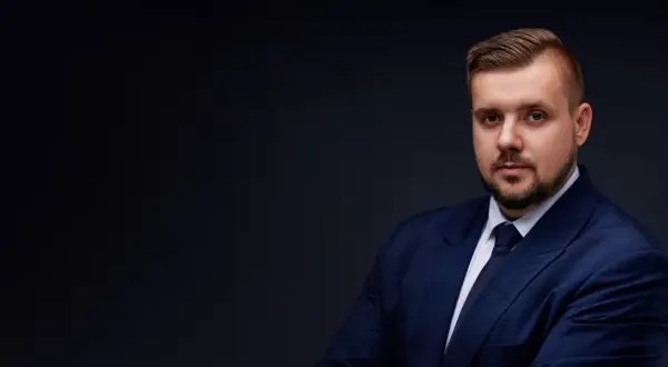 Mężczyzna w granatowym garniturze i krawacie, z zarostem, na ciemnym tle.
