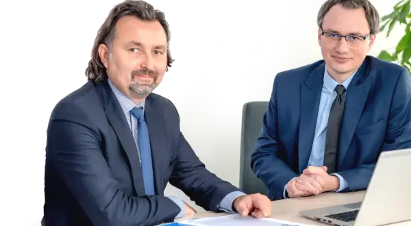 Dwóch mężczyzn w garniturach siedzi przy biurku z dokumentami i laptopem.