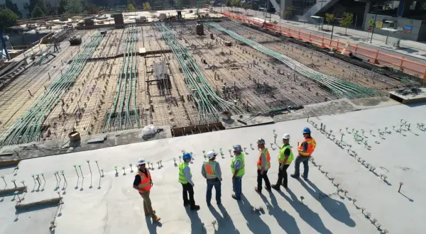 Grupa budowlańców w kaskach i kamizelkach omawia postęp prac na budowie. W tle widać zbrojenie i instalacje, zgodne z art. 62 prawa budowlanego.