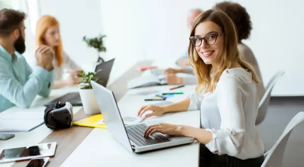 Uśmiechnięta pracownica samorządowa w okularach pracuje przy laptopie w nowoczesnym biurze.