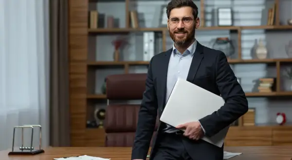 Uśmiechnięty dyrektor szkoły w garniturze, trzymający laptopa, stoi w swoim gabinecie.