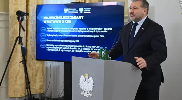 Mężczyzna przemawia przy mównicy z godłem Polski. Na ekranie za nim widnieje napis "Najważniejsze zmiany w ustawie o KRS", omawiający reformę neo KRS.