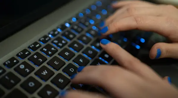 Dłonie z niebieskim lakierem na paznokciach piszą na podświetlanej klawiaturze MacBooka. Widać klawisze "twarda spacja mac".