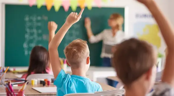 Uczniowie z podniesionymi rękami, gotowi do odpowiedzi. Wychowawca stoi przy tablicy, tłumacząc lekcję.