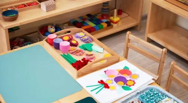 Zestaw do tworzenia kwiatów metodą Montessori: kolorowe papierowe foremki, listki, kółka i wstążki na drewnianej tacy.