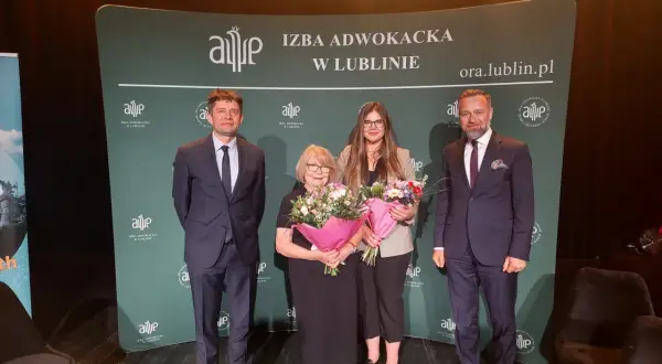 Cztery osoby w garniturach i sukience stoją na scenie z bukietami kwiatów przed zielonym banerem.
