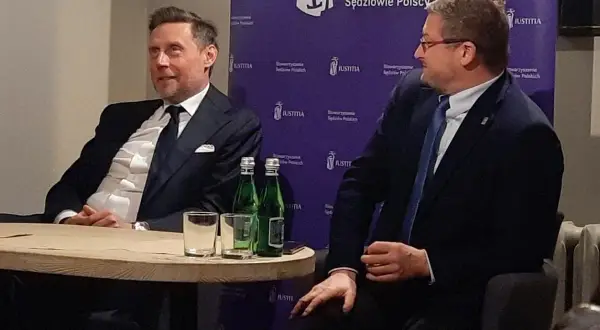 Dwóch mężczyzn w garniturach siedzi przy stole, za nimi fioletowe tło z logo "JUSTITIA".