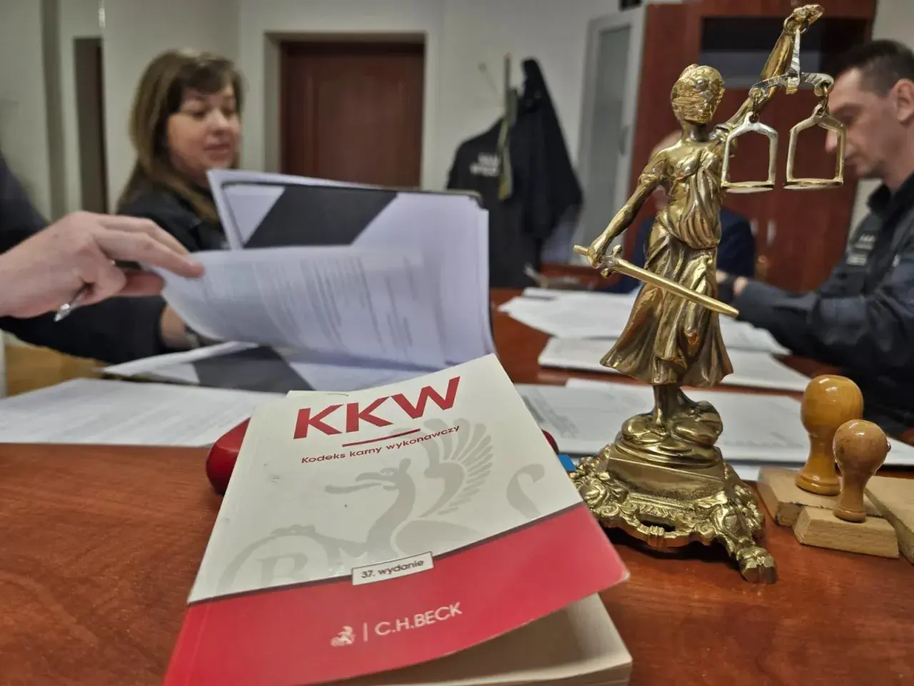 Na biurku leży Kodeks karny wykonawczy, obok Statua Temidy. W tle widać osoby pracujące nad dokumentami, co może sugerować sprawy związane z dozorem elektronicznym.