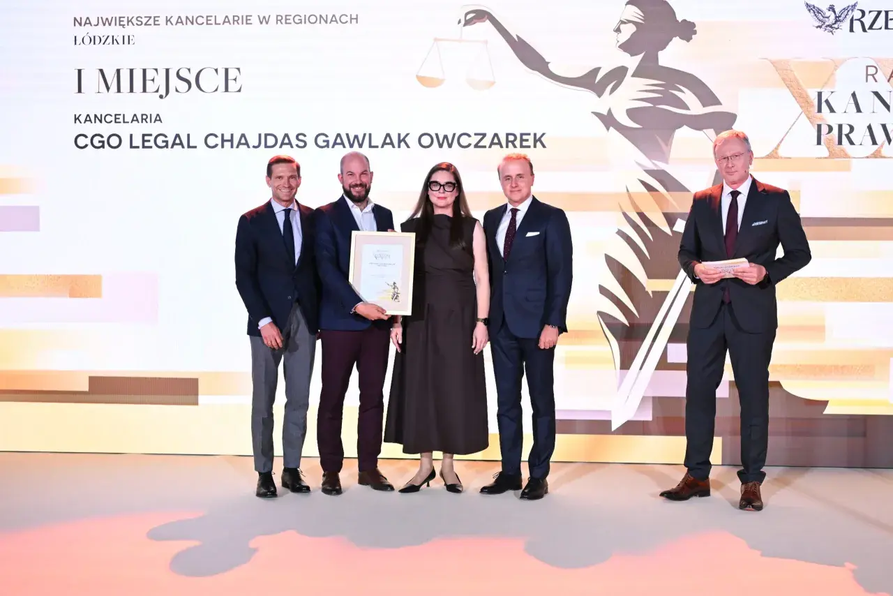 Pięć osób w strojach formalnych pozuje do zdjęcia na tle sceny z grafiką i napisem "I MIEJSCE".