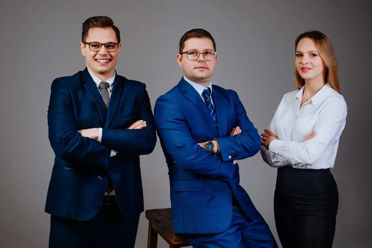 Trzy osoby w garniturach i białej koszuli, stojące z rękami skrzyżowanymi na piersiach.