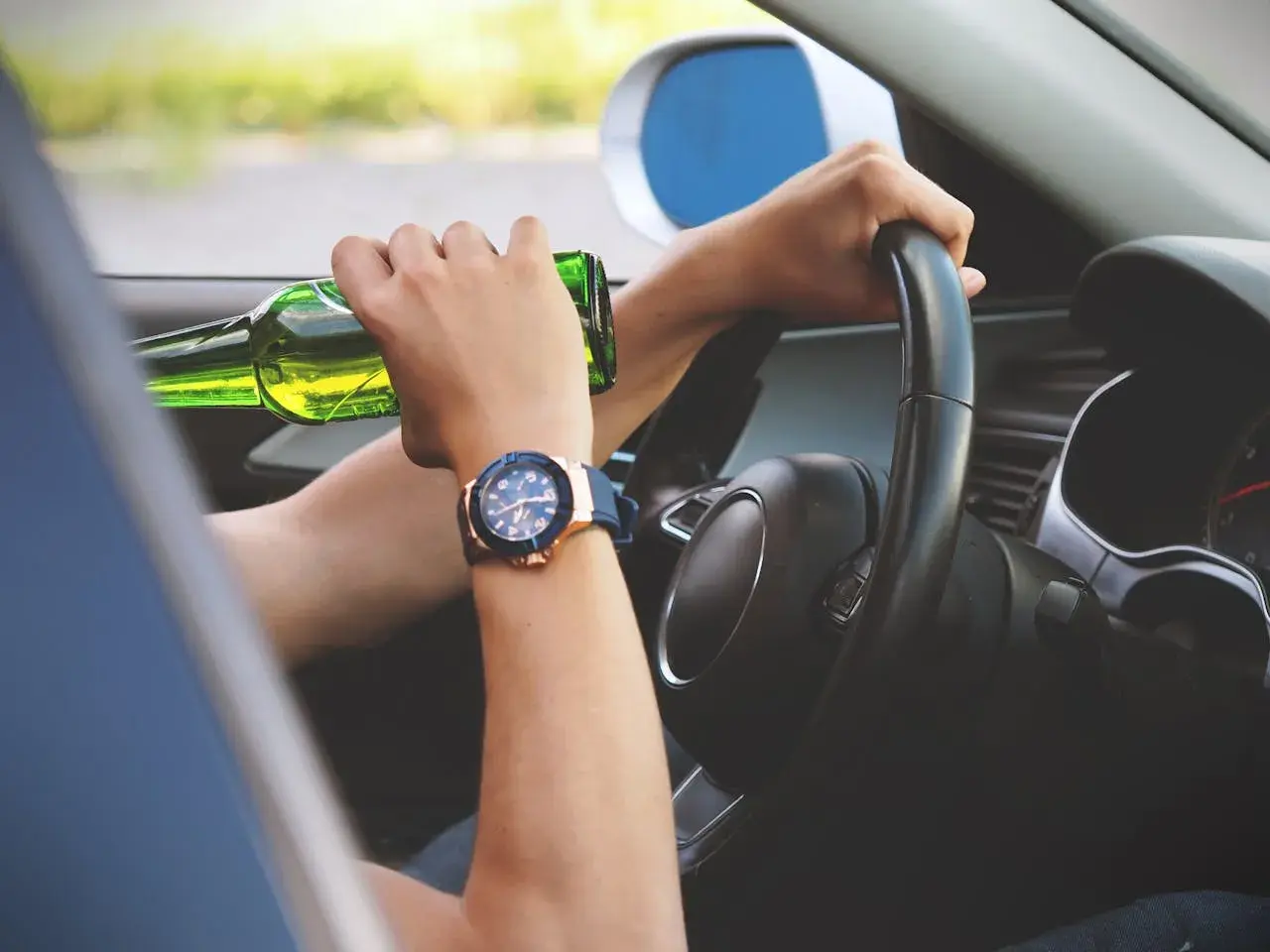 Ręce na kierownicy, jedna trzyma butelkę piwa. Ryzykowne zachowanie, stan po użyciu alkoholu.