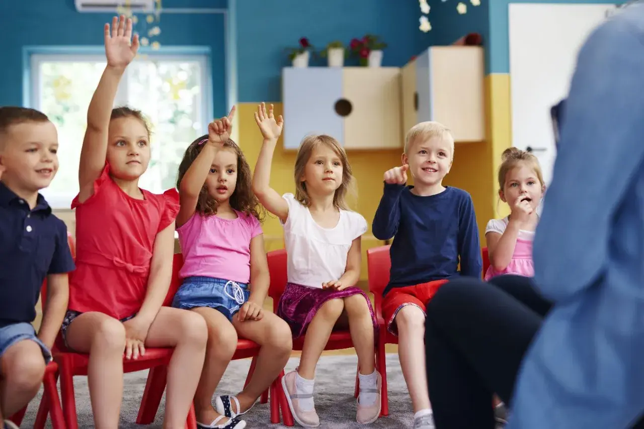 Dzieci na czerwonych krzesłach podnoszą ręce, jakby odpowiadały na pytanie, czy zerówka jest obowiązkowa.