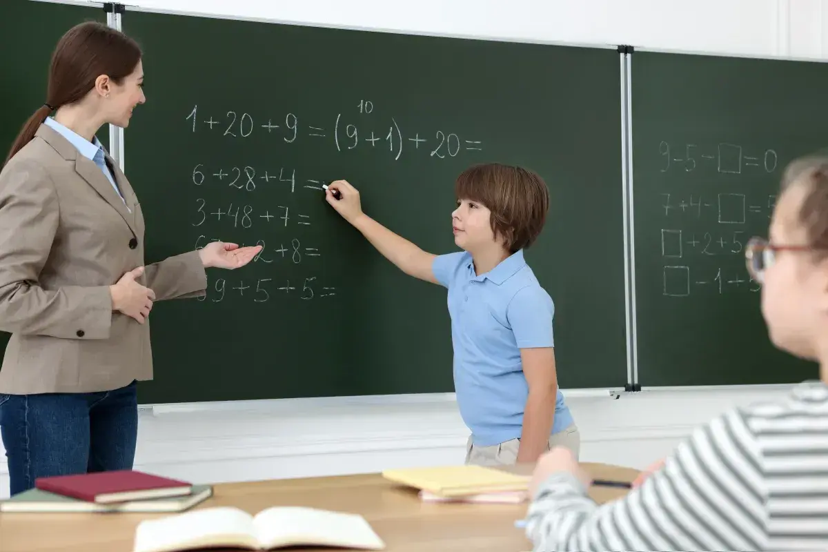 Nauczycielka pomaga uczniowi rozwiązać zadania matematyczne przy tablicy. Dzieci uczą się, wypełniając swój obowiązek szkolny.