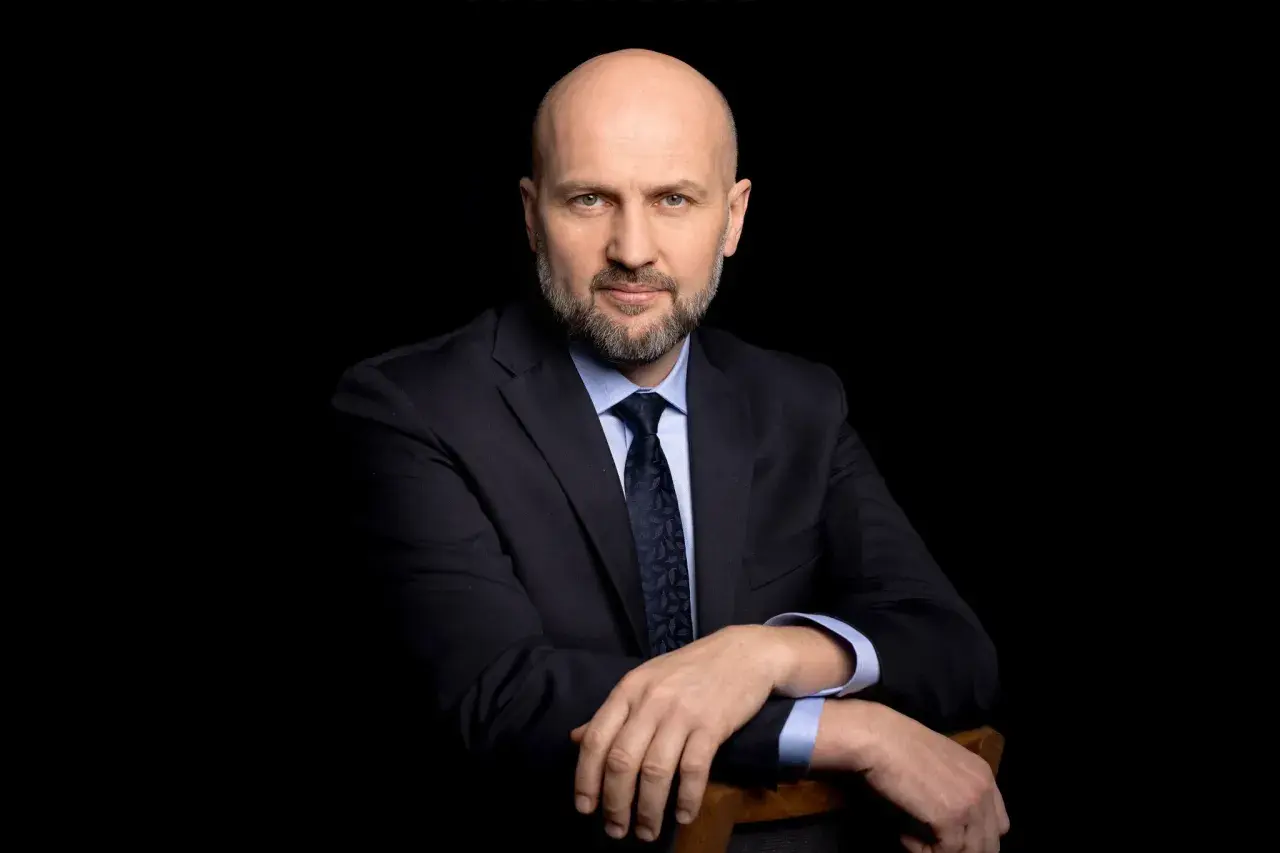 Łysy mężczyzna w garniturze i krawacie, oparty o krzesło, na czarnym tle.