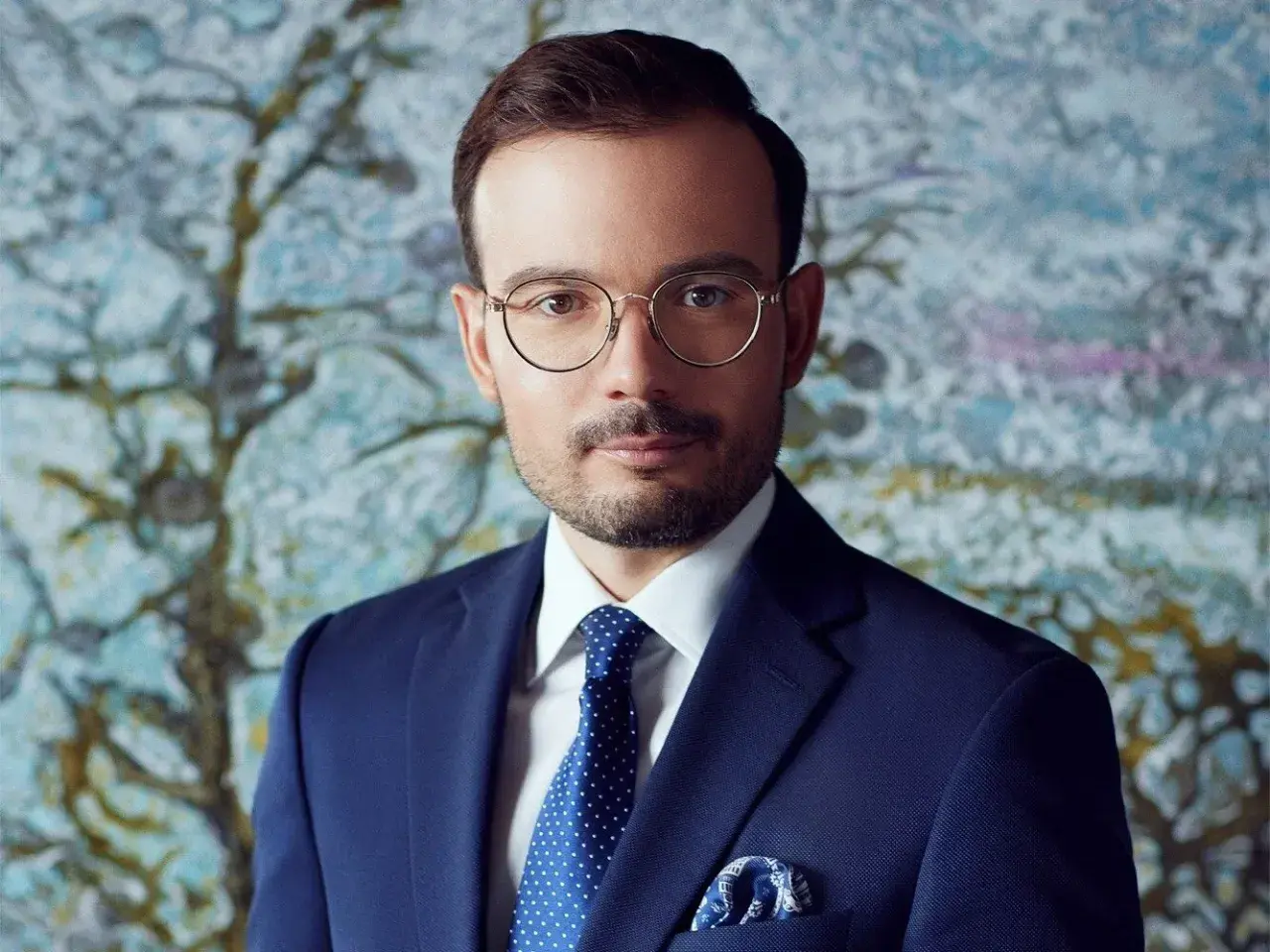 Mężczyzna w okularach i granatowym garniturze na tle abstrakcyjnego obrazu.
