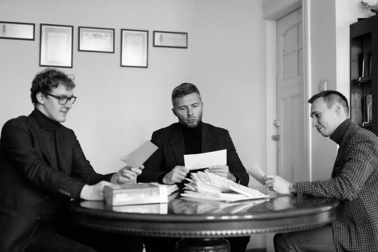 Trzech mężczyzn przy okrągłym stole analizuje dokumenty. Jeden z nich trzyma kopertę, jakby właśnie otrzymał wyrok w zawieszeniu.