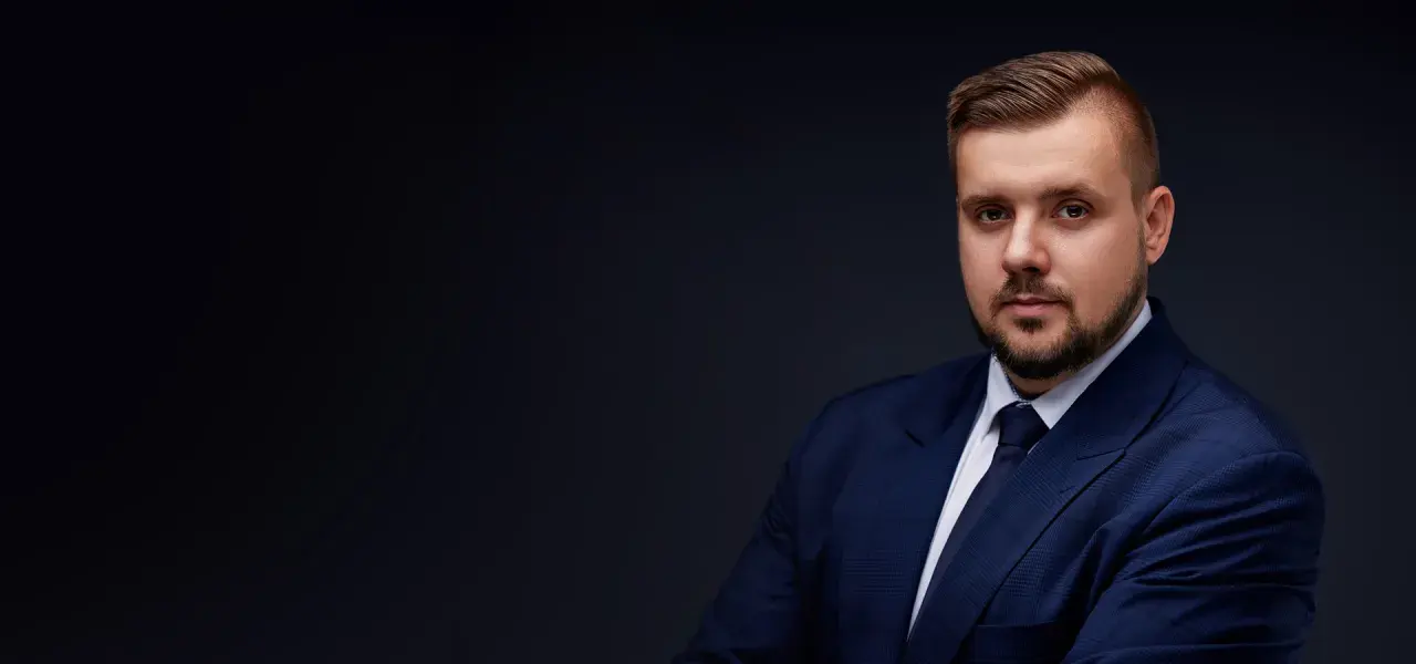 Mężczyzna w granatowym garniturze i krawacie, z zarostem, na ciemnym tle.