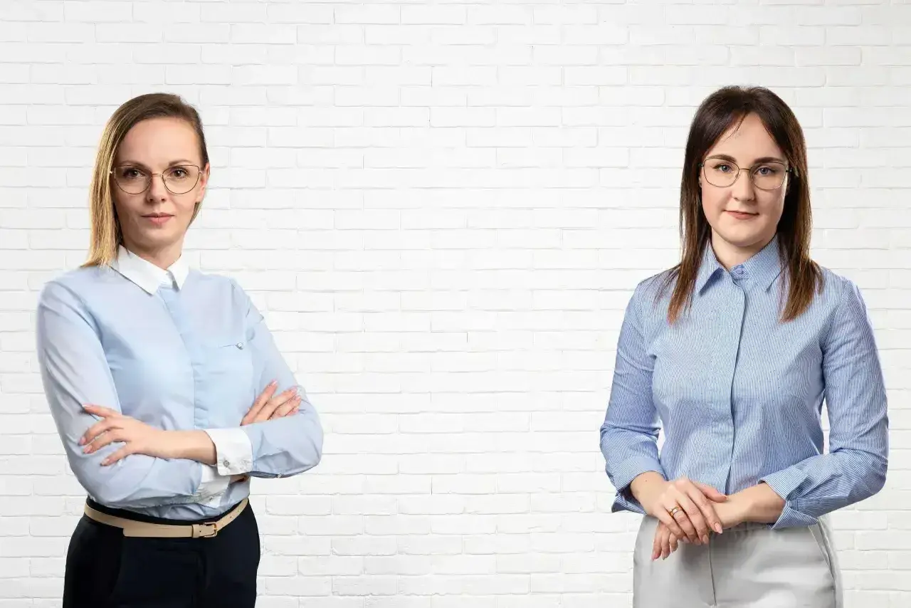 Dwie kobiety w koszulach i okularach na tle białej ceglanej ściany.