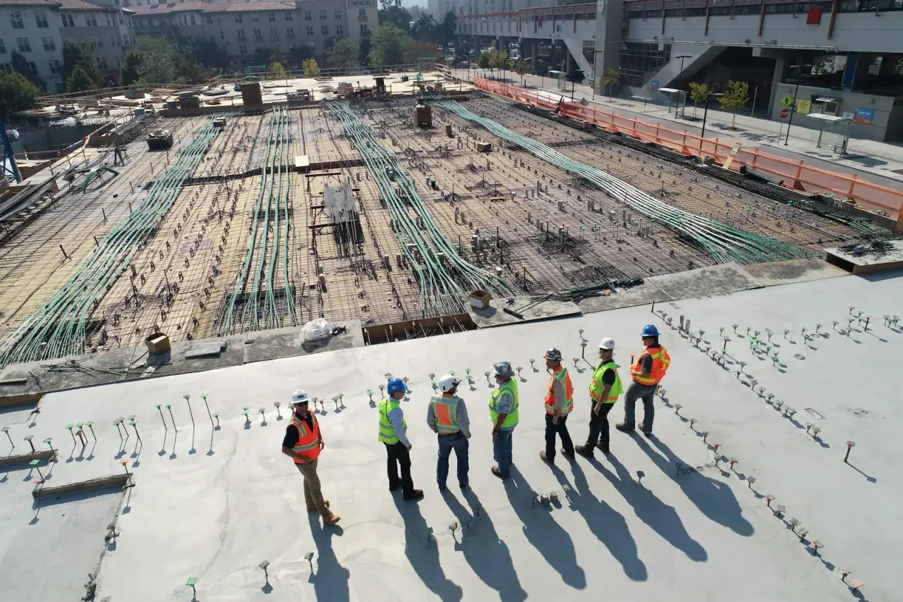 Grupa budowlańców w kaskach i kamizelkach omawia postęp prac na budowie. W tle widać zbrojenie i instalacje, zgodne z art. 62 prawa budowlanego.