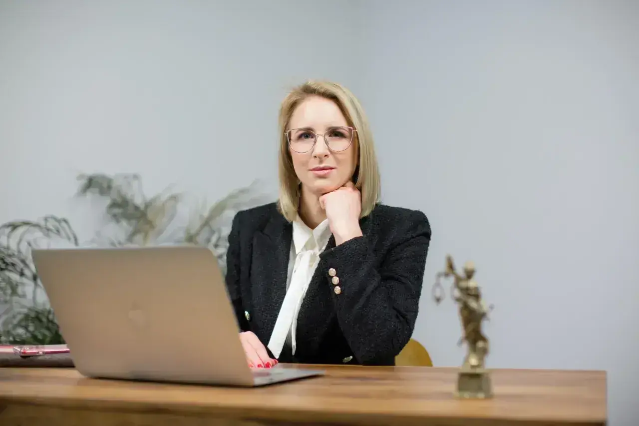 Kobieta w okularach i żakiecie siedzi przy biurku z laptopem i statuetką Temidy.