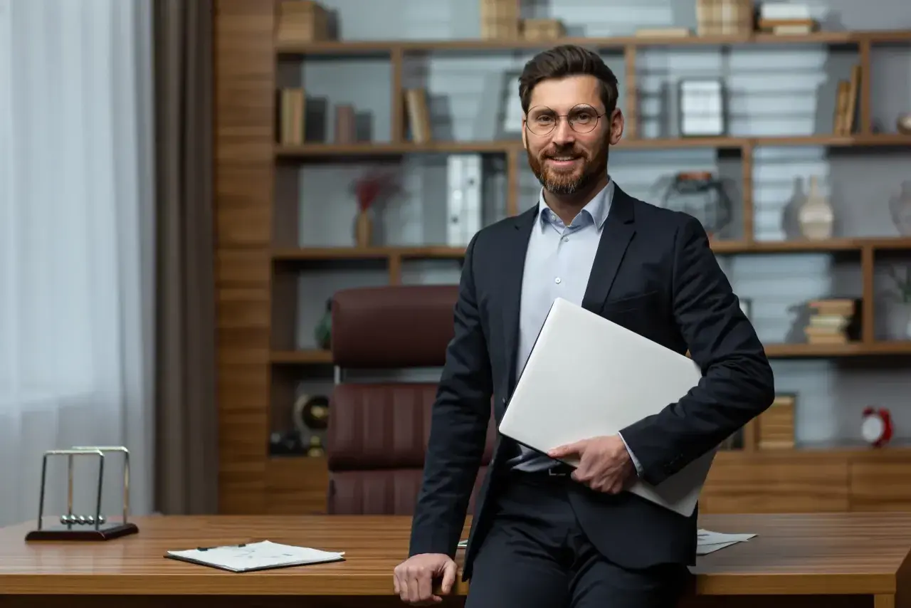 Uśmiechnięty dyrektor szkoły w garniturze, trzymający laptopa, stoi w swoim gabinecie.