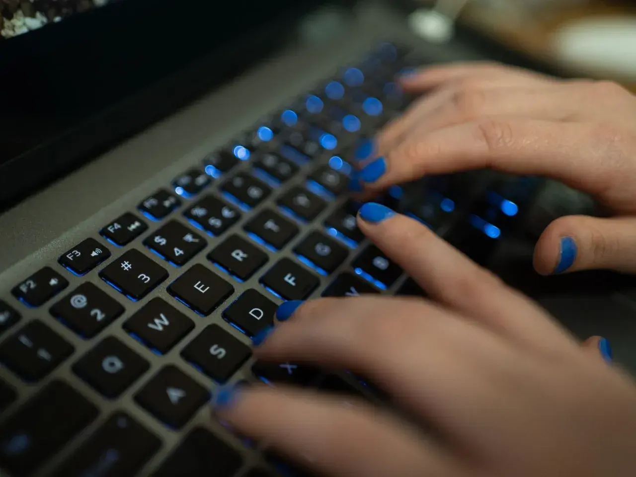Dłonie z niebieskim lakierem na paznokciach piszą na podświetlanej klawiaturze MacBooka. Widać klawisze "twarda spacja mac".