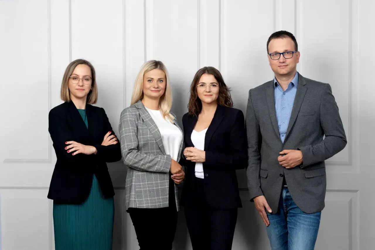 Cztery osoby w strojach biznesowych stoją obok siebie na jasnym tle.