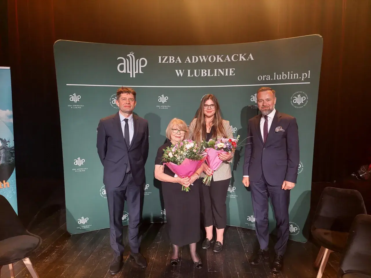 Cztery osoby w garniturach i sukience stoją na scenie z bukietami kwiatów przed zielonym banerem.