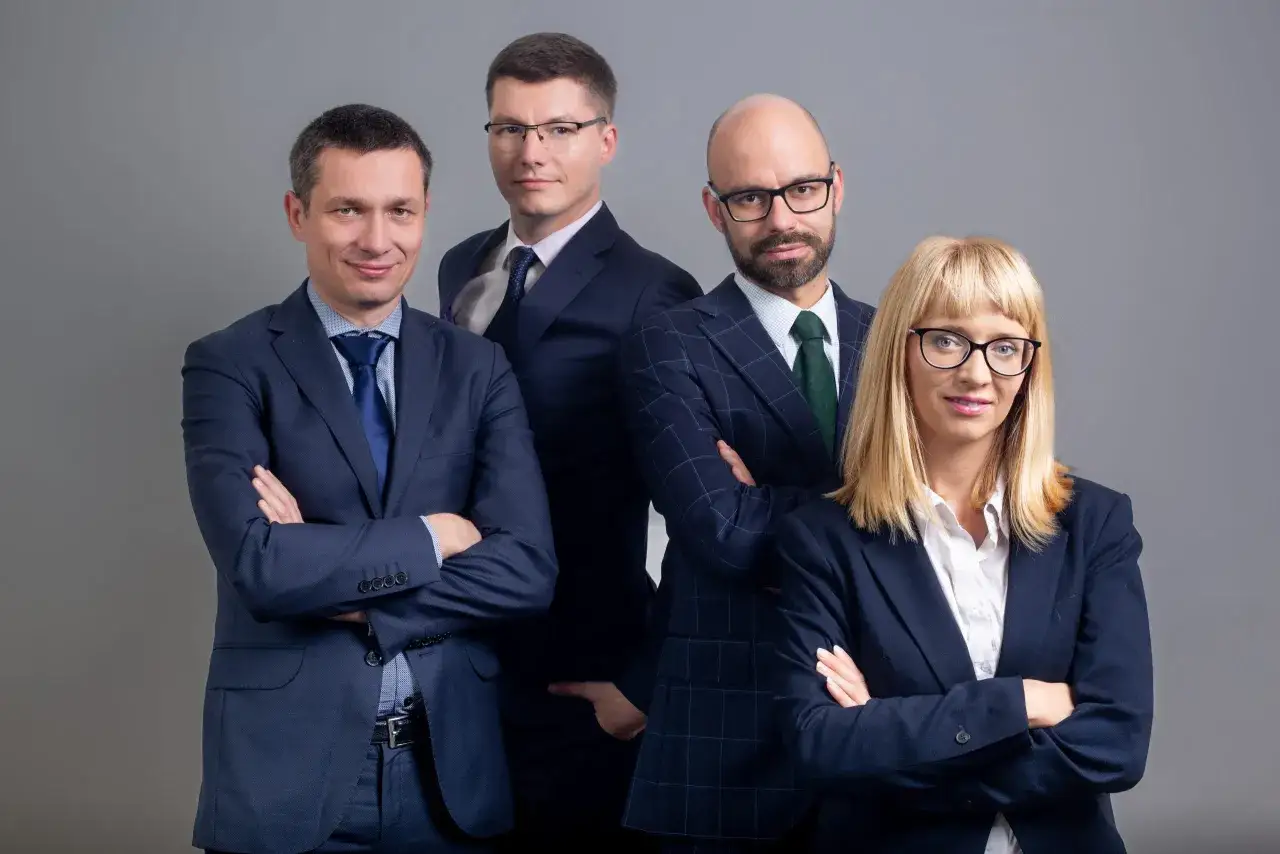 Cztery osoby w garniturach, stojące na szarym tle, z rękami skrzyżowanymi na piersi.