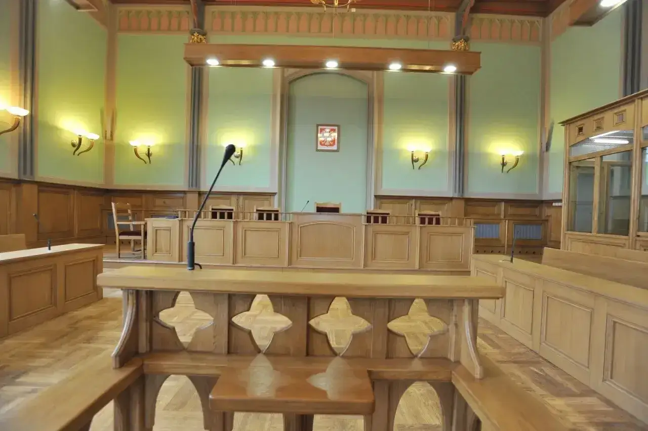 Drewniana sala sądowa, gotowa na proces sądowy. W centrum widoczny pulpit z mikrofonem, a za nim ławy dla sędziów i urzędników.