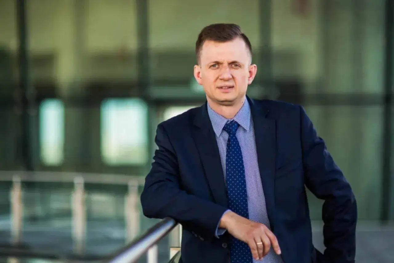 Mężczyzna w garniturze i krawacie opiera się o barierkę.