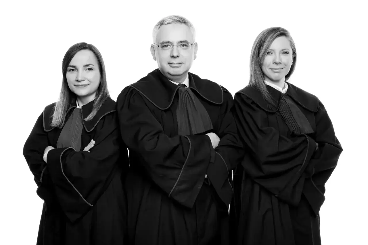 Trzy osoby w togach prawniczych, dwie kobiety i mężczyzna, na białym tle.