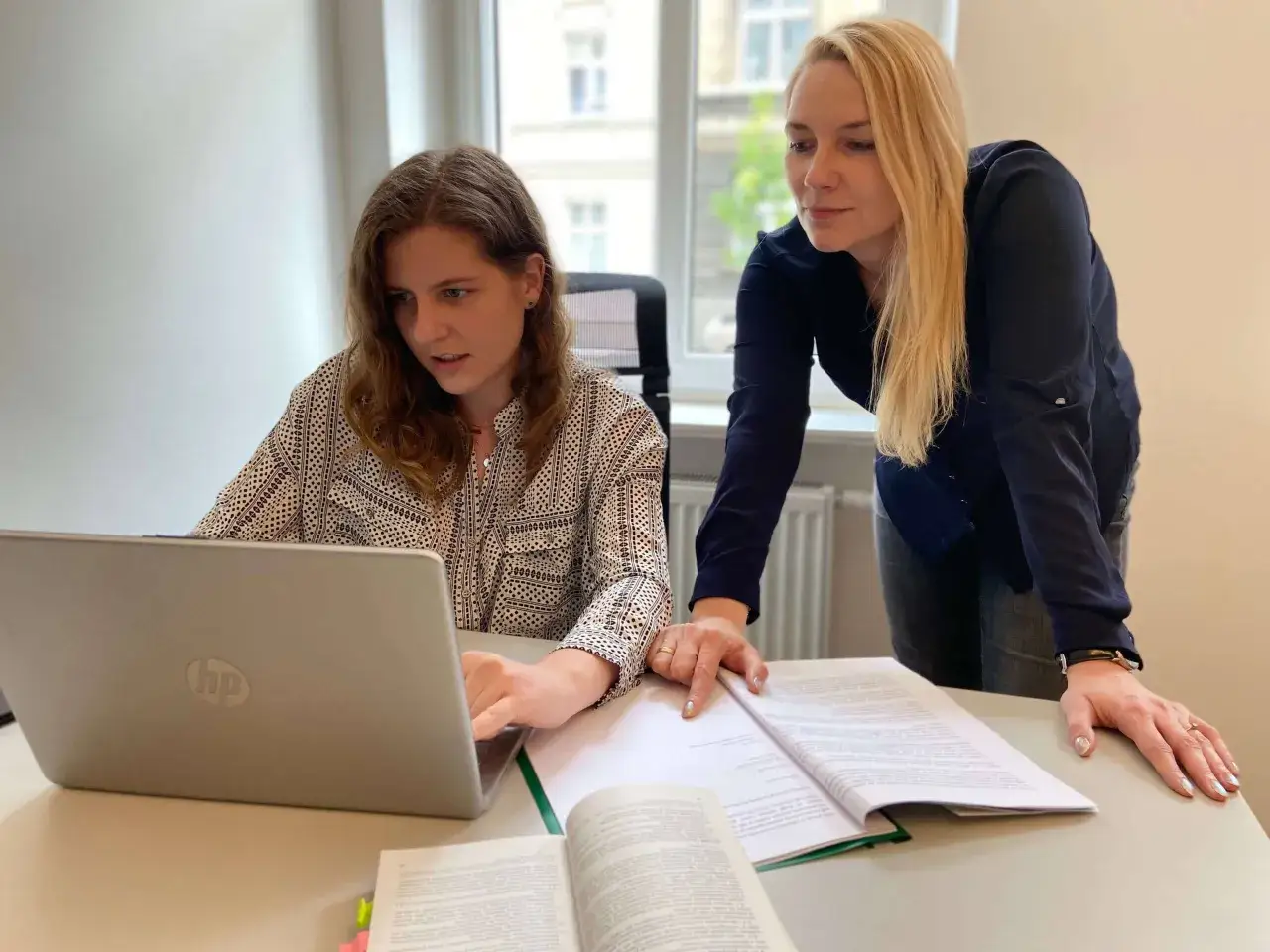 Dwie kobiety analizują dokumenty na biurku. Jedna pracuje przy laptopie, druga wskazuje palcem na otwartą księgę. Być może przygotowują się do formalności związanych z rozwodem u notariusza.