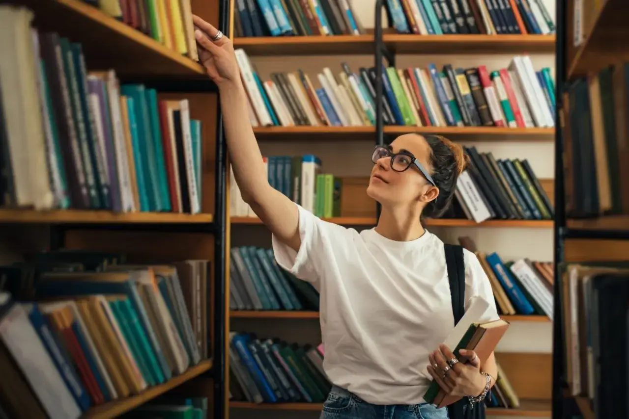 Dziewczyna w okularach szuka książki na półce w bibliotece. To jej profil w liceum, pełen nauki i odkryć.