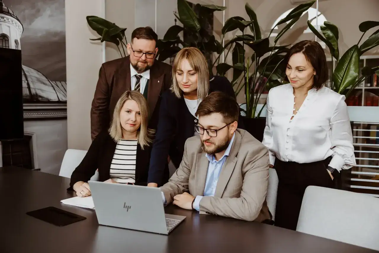 Zespół prawników przy stole z laptopem, w tle rośliny i grafika budynku.