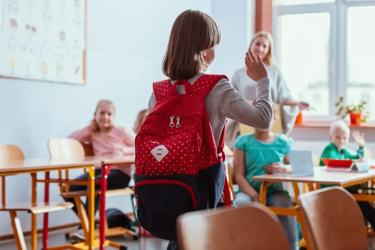 Dziewczynka z plecakiem wchodzi do klasy, czując niepokój związany z fobią szkolną. Nauczycielka i inni uczniowie witają ją.