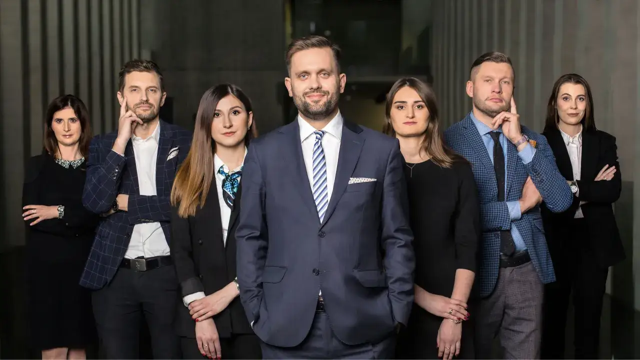Grupa prawników w garniturach i kostiumach stoi przed betonową ścianą.