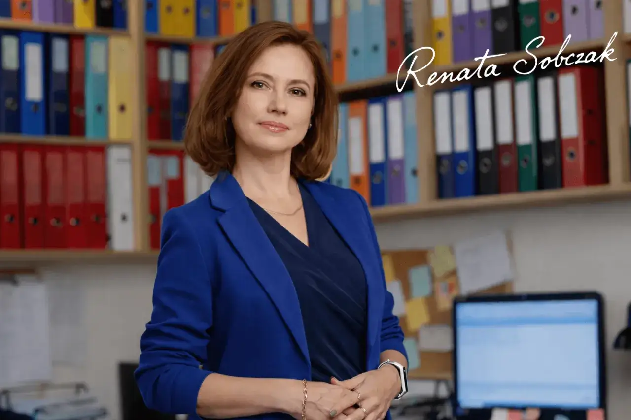 Kobieta w niebieskiej marynarce w biurze, z półkami pełnymi segregatorów w tle.